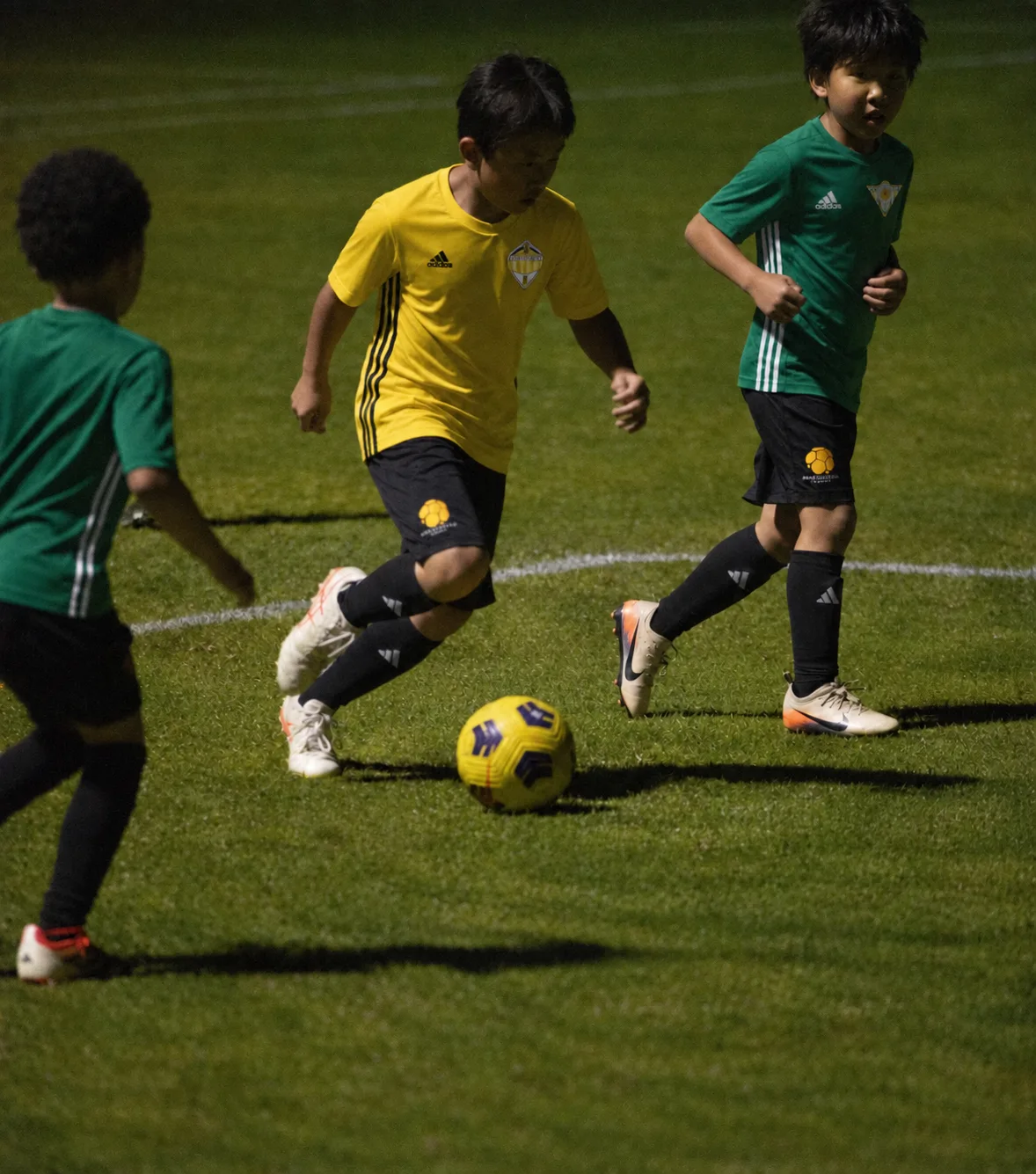 Three young Apex players chasing the ball across floodlit grass, one in a yellow shirt running on to it